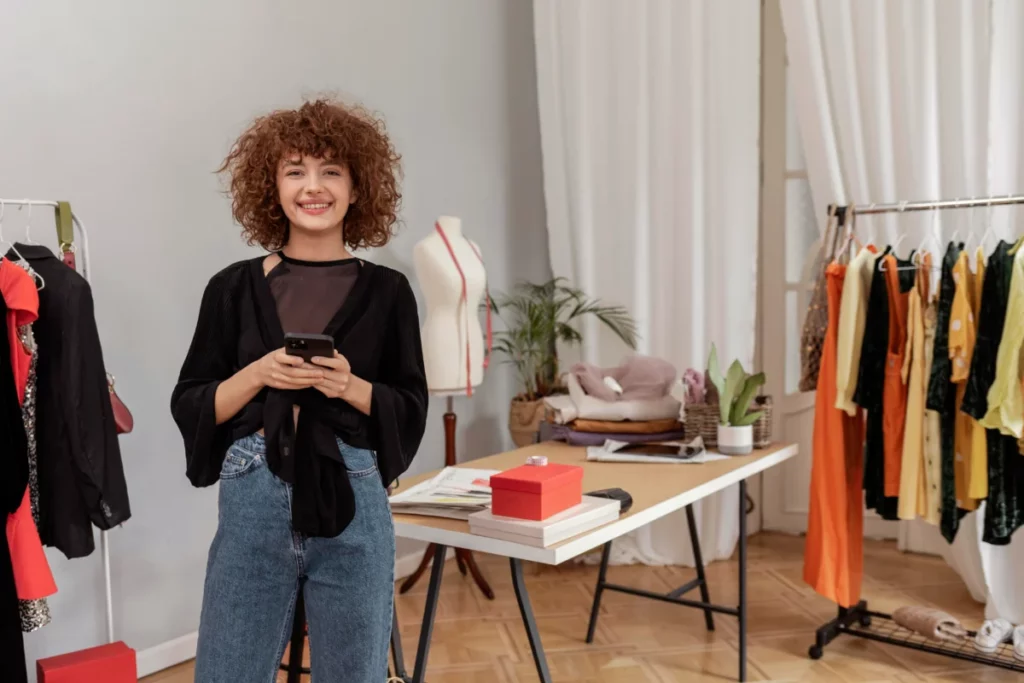 mulher designer de roupas em ateliê com celular na mão - como começar a vender roupas do zero