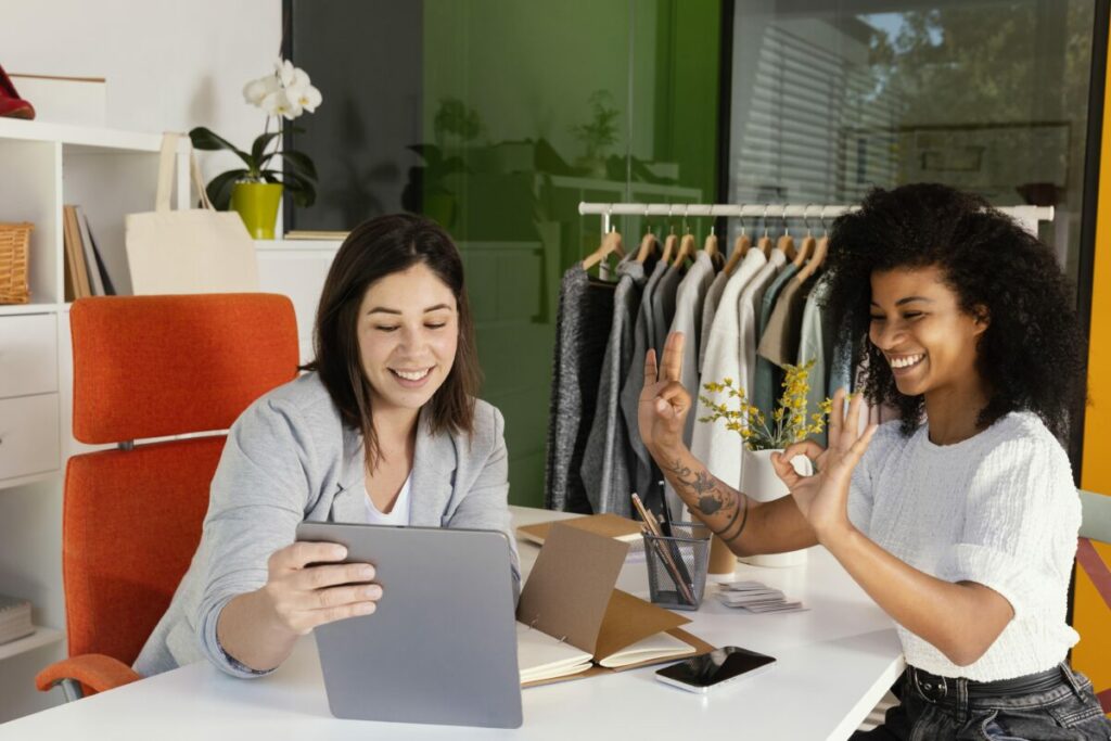 mulheres sentadas em lados opostos de uma mesa, olhando para um tablet com comércio ao fundo - digital commerce