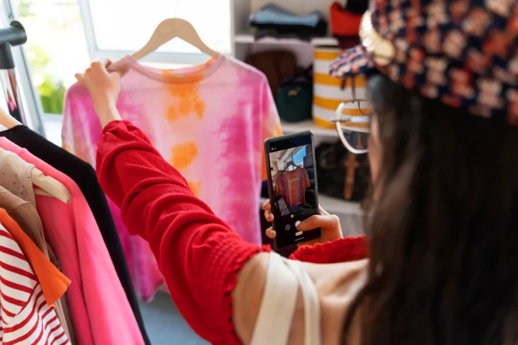 vista de trás de uma mulher segurando uma peça de roupa com uma mão e tirando foto com a outra - como começar a vender roupas do zero