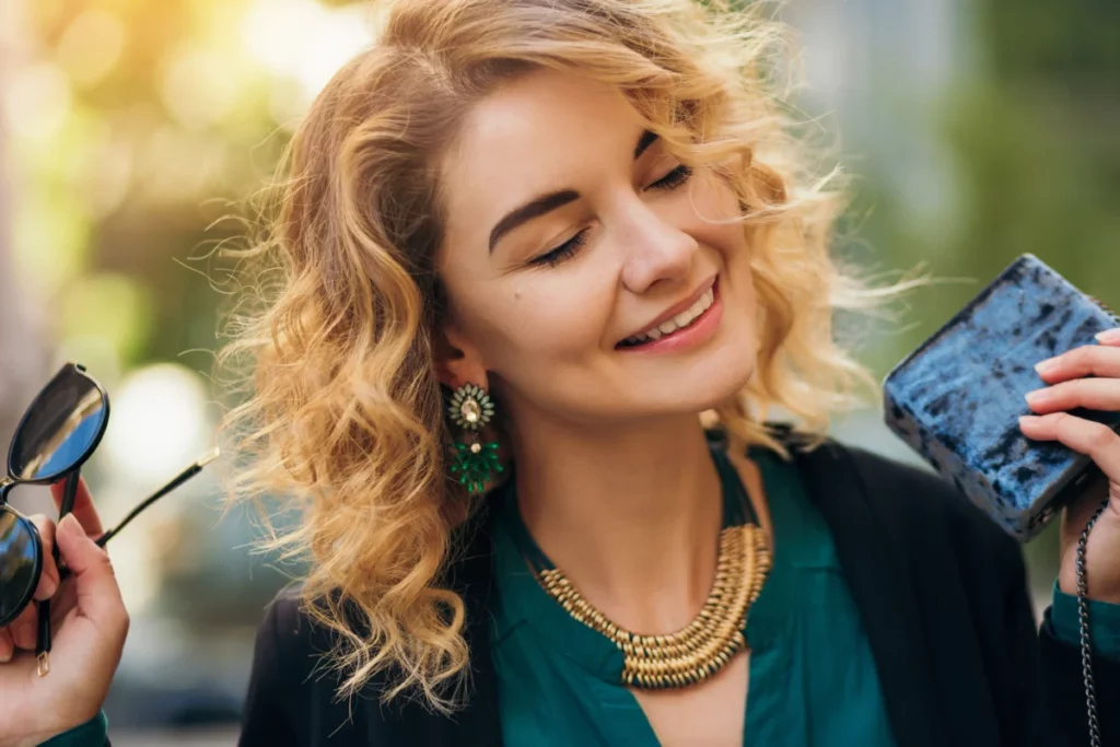 mulher estilosa segurando óculos de sol e cartira, sorrindo ao usar colar e brincos - ideias de acessórios para vender