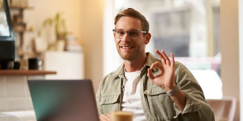 homem-de-negocios-usando-oculos-e-sorrindo-ao-mostra-um-sinal-de-ok-trabalhando-no-computador-em-um-cafe-como-fazer-uma-loja-virtual