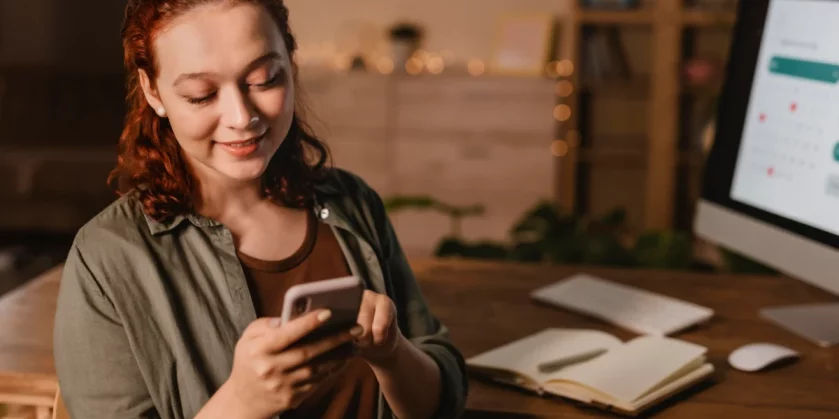 mulher-sorrindo-mexendo-no-celular-em-frente-ao-computador-atendimento-via-whatsapp